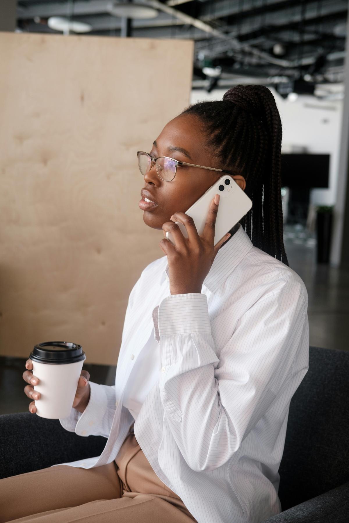 Vrouw in witte blouse die haar telefoon aan haar oor houdt en in de rechterhand een koffie to go vasthoudt